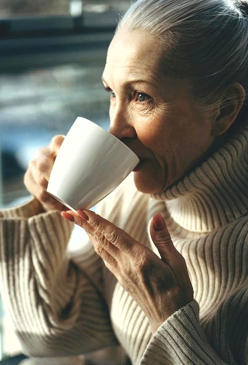 A woman in her eighties drinking coffee by a window.