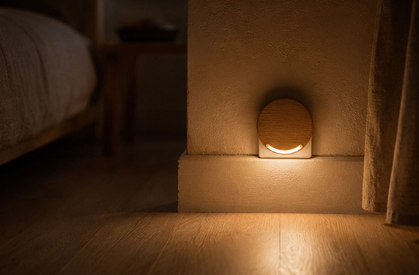 A round oak night light glowing at skirting height beside a bed.